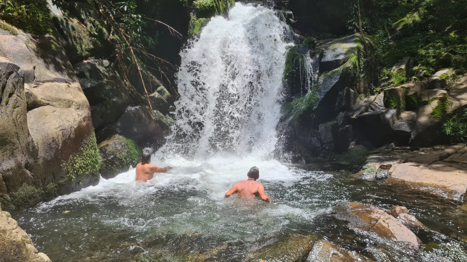 Two people bathing beneath a waterfall among rocks in the tropical rainforest.